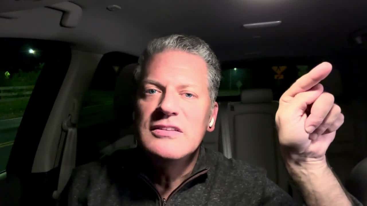 A man with grey hair and wearing black is in the back of a car.
