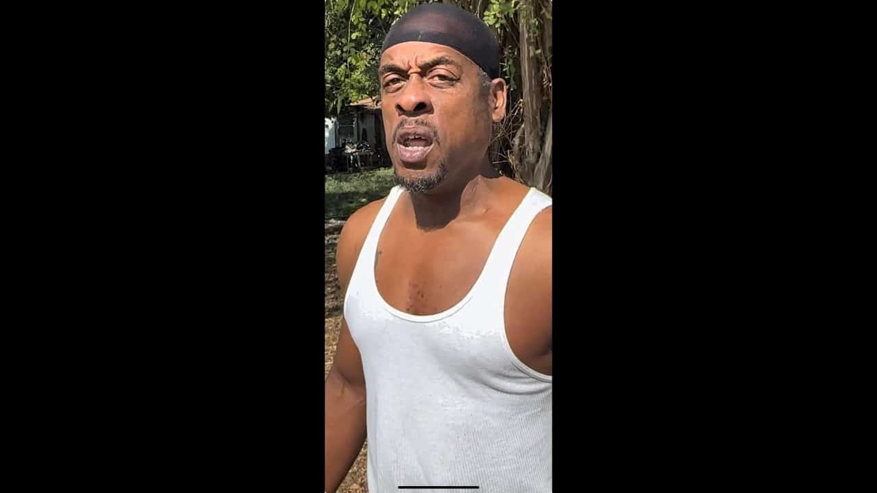 A man in white shirt and black hat standing next to tree.
